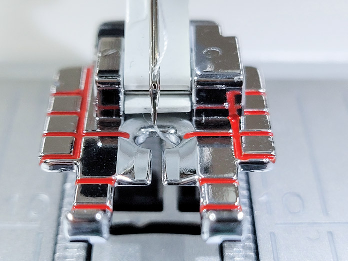A needle in the center of a metal presser foot with red markings; Husqvarna Viking Tribute 150C