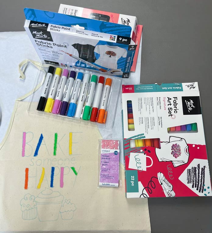 A box of Mont Marte Fabric Paint Sticks with the paint sticks shown, a box of Mont Marte Fabric Art Set and a package of UNIQUE Rick Rack are set next to the apron with the design on the apron partially painted inProgressing with the coloring with the paint sticks, , Fabric Fun Apron for Embellishment