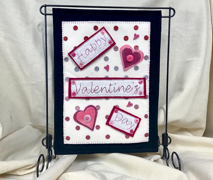 A pink, red, cream and black Valentine’s Day banner hangs from a black metal stand on a cream-colored background. CRAFTING ESSENTIALS Bottle of Buttons - Pink Tones - 75g (2.6oz), HEATNBOND EZ Print Feather Lite 10 pcs - 22 x 28cm (81⁄2″ x 11″), SULKY Cotton Petites 6 Spool Thread Set - Rosewood Manor Assortment, UNIQUE Hook & Loop Sew-On Tape - 19mm x 10m (3⁄4″ x 11yd) - Black, OLFA RTY-2/DX - Deluxe Ergonomic Handle Rotary Cutter 45mm, OMNIGRID Ruler - 6″ x 12″, ELAN Novelty 2-Hole Button - Rose - 18mm (3⁄4″) - Heart - 3 count, UNIQUE SEWING Chenille Needles - sizes 18/24 - 6pcs, ODIF OdiShine Glitter Gel, DMC Embroidery Tracing/Transfer Paper, UNIQUE QUILTING Clever Clips Small - 12 pcs, free tutorial.
