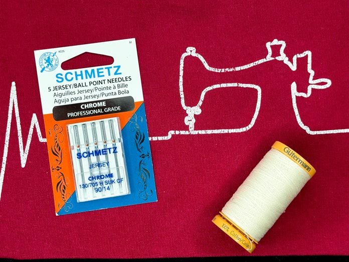 SCHMETZ Chrome Jersey five pack of needles and a spool of ivory colored GÜTERMANN Cotton 50wt thread on a red T-shirt with a motif of a sewing machine with a heartbeat.