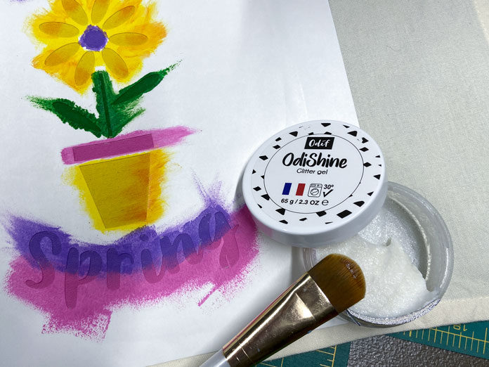 A small jar of Odishine glitter gel is shown with its lid removed, on top of a tea towel with a stencil ironed to it. The stencil has already been painted with pink purple and yellow paint. A paintbrush is laying on top; Sew Easy Freezer Paper for Quilting and Applique, Mont Marte Signature Fabric Paints, Odif OdiShine Glitter gel, Odif OdiShine Glitter Gel – Opal 65g