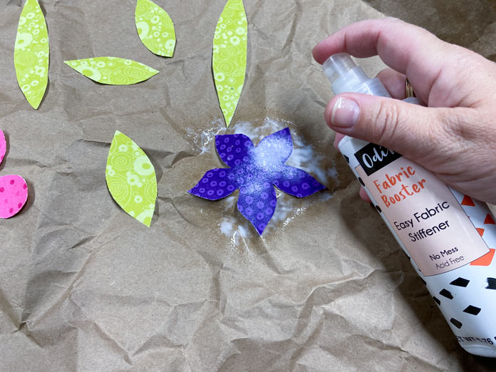 A hand is shown spraying a white liquid onto a purple fabric. Five green leaves and the edge of a pink flower can also be seen. A crumpled piece of brown paper is the background; Odif Fabric Booster
