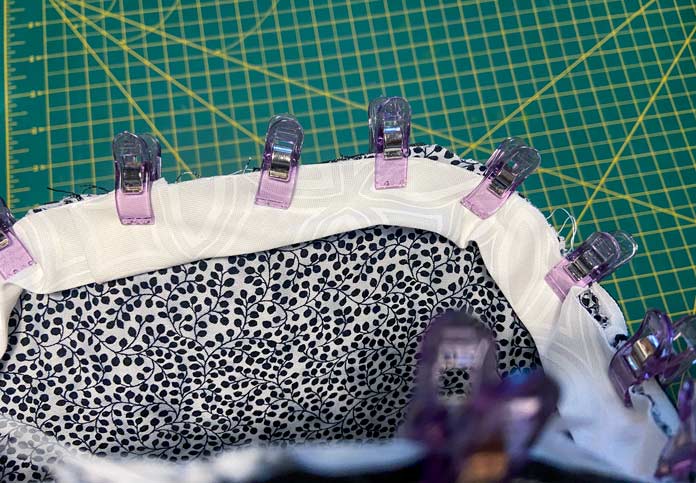 Small purple clips hold white binding to the inside of the tote bag along the top edge of the black and white lining fabric. A green cutting mat can be seen in the background.