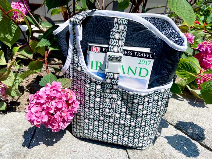 A multicolored crossbody bag sits on a garden wall in front of a pink hydrangea bush. A 2017 Ireland travel guide can be seen in the front pocket. There is a strap and buckle holding the book in place.