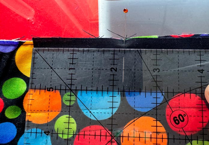 An OLFA ruler measures 2¼” from the end of a black fabric strip sewn to a quilt backing of black fabric with brightly colored circles on it while a pin is placed through the fabric strip at the 2¼” mark. A red sewing table is in the background. Hemline Gold Multi-Use Craft Bag (28.5cm x 36.5cm x 17.8cm), Hemline Gold Sewing Kit, Hemline Gold Quilters 4-in-1 Multi-Mat (30cm x 24cm), Hemline Gold Quilters Clips (Pack of 30), Hemline Gold Retractable Tape Measure - 150cm/60, oliso M3Pro Project Iron – Coral, OLFA 45mm RTY-2/DX/MAG Ergonomic Rotary Cutter – Magenta, OLFA RM-MG - 24″ x 36″ Double Sided Rotary Mat, UNIQUE Sewing Tools Holder, OLFA QR-6x12 - 6″ x 12″ Frosted Acrylic Ruler, OLFA QR-6x24 - 6″ x 24″ Frosted Acrylic Ruler, Fairfield Quilter's 80/20 Quilt Batting - 229 x 274cm (90″ x 108″) 