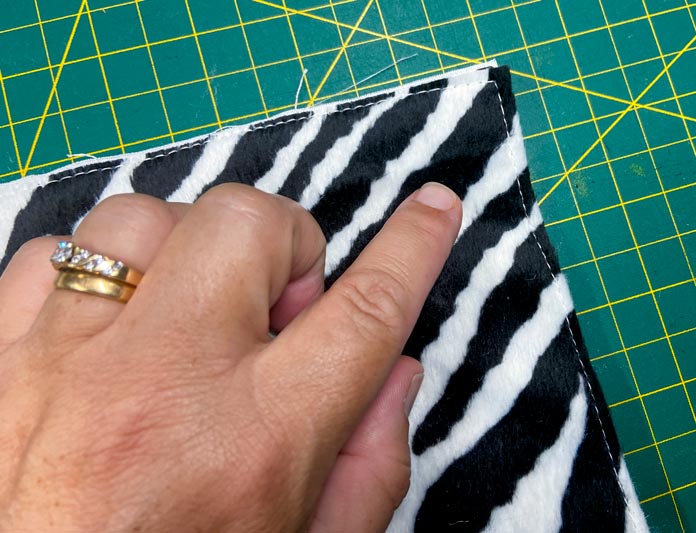 A hand is shown pointing to the basting stitches on a black and white piece of faux zebra fur on top of a green cutting mat. Oliso M2Pro Mini Project Iron, FAIRFIELD Low-Loft Quilt Batting - Crib - 114 x 152cm (45″ x 60″), UNIQUE QUILTING Clever Clips, OLFA CHN-1 - Chenille / Textile Cutter, UNIQUE CREATIV Trim Pack - Black & White, FABRIC CREATIONS Burlap Fabric - Oyster - 45 x 61cm (18″ x 24″), FABRIC CREATIONS Faux Fur Fabric - Zebra - 45 x 76cm (18″ x 30″), UNIQUE SEWING Double Adjust Parachute Buckle - Plastic - 25mm (1″) White, CRAFTING ESSENTIALS Bottle of Buttons - Pink Tones - 75g (2.6oz), UNIQUE SEWING Invisible Magnetic Snaps - Sew-On - 12mm (1⁄2″) - 2 sets, UNIQUE QUILTING Stitch-N-Steam - 157 x 45cm (62″ x 18″), FABRIC CREATIONS Fabric Bundle (5pcs) - Metallic Gold - 45 x 53cm (18in x 21in), Oliso Pro TG1600 Pro Plus Smart Iron, UNIQUE QUILTING Clever Clips Large - 12pcs, sewing tutorial 