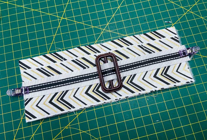 A brown buckle with black and white webbing threaded through it is clipped to a quilt sandwich made of white, black, and gold fabric. All lay on top of a green cutting mat. OLFA CHN-1 - Chenille / Textile Cutter, UNIQUE QUILTING Stitch-N-Steam - 157 x 45cm (62″ x 18″), FABRIC CREATIONS Fabric Bundle (5pcs) - Metallic Gold - 45 x 53cm (18in x 21in), UNIQUE SEWING Self-Gripping Fastener Strip - 25cm (10″) - White, UNIQUE Webbing 25mm x 10m (1″ x 11yd) - White, an ELAN Trench Buckle - 45mm (13⁄4″) - Brown, UNIQUE CREATIV Trim Pack - Black & White, UNIQUE QUILTING Clever Clips Small - 12 pcs, CLOVER 7807 - Hold it Precision Stiletto, UNIQUE SEWING Sewing Tools Holder, UNIQUE QUILTING Wool Pressing Mat - 18″ x 24″ - Grey, OLISO M2Pro Mini Project Iron with Solemate, UNIQUE QUILTING Clever Clips Large - 12pcs, UNIQUE SEWING Invisible Magnetic Snaps - Sew-On - 12mm (1⁄2″) - 2 sets, COSTUMAKERS Activewear One Way Separating Zipper 23cm (9″) - White - 1760, UNIQUE SEWING Double Adjust Parachute Buckle - Plastic - 25mm (1″) - White, free tutorial