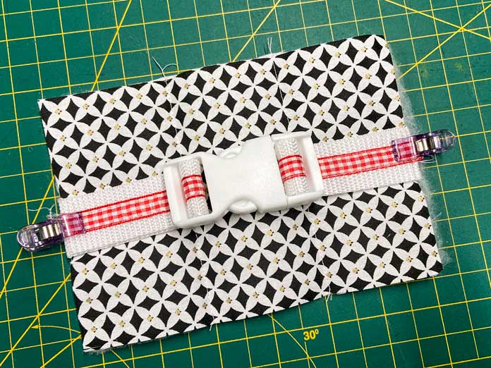 Small purple clips hold together a black and white quilt block and the ends of white webbing with a red gingham ribbon sewn down the center. The webbing is threaded through a white parachute buckle. A green cutting mat can be seen in the background. UNIQUE SEWING Self-Gripping Fastener Strip - 25cm (10″) - White, UNIQUE Webbing 25mm x 10m (1″ x 11yd) - White, ELAN Trench Buckle - 45mm (13⁄4″) - Brown, UNIQUE CREATIV Trim Pack - Black & White, UNIQUE SEWING Invisible Magnetic Snaps - Sew-On - 12mm (1⁄2″) - 2 sets,  SOFTKUT Pinking Shears (Scissors) - 81⁄2″ (21.6cm), COSTUMAKERS General Purpose Closed End Zipper 20cm (8″) - White - 1700, UNIQUE CREATIV Trim Pack - Red & Green, OLFA RTY-2/DX - Deluxe Ergonomic Handle Rotary Cutter 45mm, UNIQUE QUILTING Clever Clips Small - 12 pcs, UNIQUE SEWING Double Adjust Parachute Buckle - Plastic - 25mm (1″) - White, CRAFTING ESSENTIALS Bottle of Buttons - Pink Tones - 75g (2.6oz), UNIQUE Braided Elastic 3mm x 3.7m - White, free tutorial