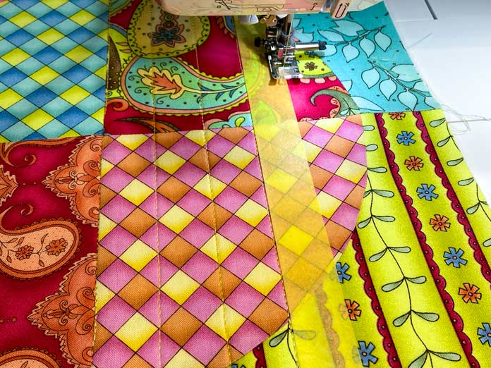 A multicolored quilt block is being machine quilted with yellow thread. A yellow strip of painter’s tape has been stuck to the fabric to use as a guide. 