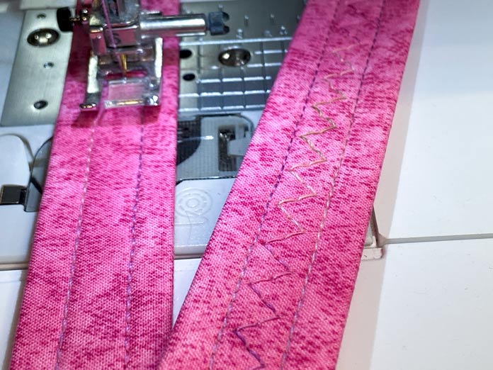 A sewing machine is shown stitching a zigzag down the middle of one of the pink fabric handles using a variegated pink thread. The second completed handle sits off to the side on top of the sewing machine bed.