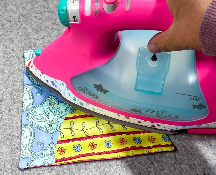 A pink and blue iron is shown pressing a multicolored quilt block on a grey wool pressing mat.