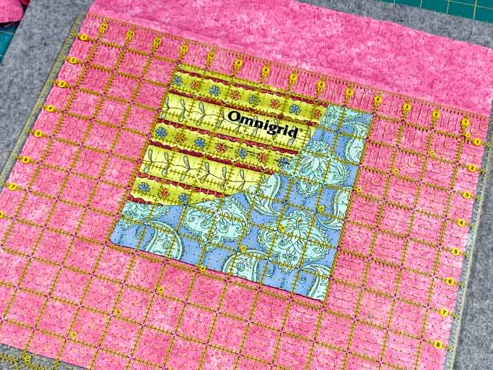 A multicolored pocket is shown on top of a large square of pink fabric. A yellow gridded rotary cutting ruler is sitting on top of both, centering the pocket.