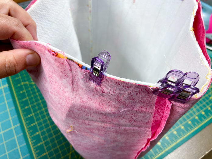 A hand holds the top edge of a tote bag. The lining of the tote bag is on the outside with its wrong side out. Purple clips secure the outside of the bag and the lining together, preparing them for sewing. A green cutting mat can be seen in the background.