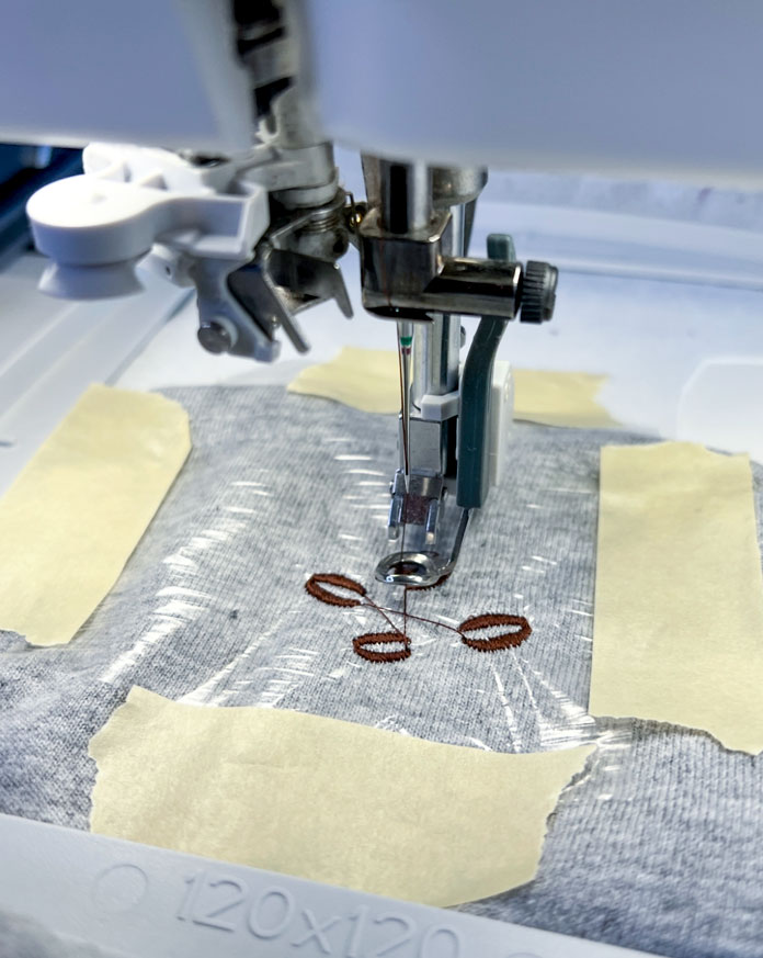 The sleeve is securely framed in a 120x120 hoop, stabilized with white sew-in interfacing at the back and covered with a layer of clear water-soluble topping. The sleeve fabric and topping are held firmly in place with masking tape at all four corners of the hoop to ensure no shifting during stitching. The embroidery is in progress. 