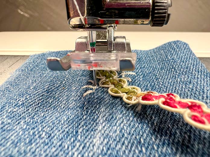 Close-up of the 2A Fancy Stitch Foot on the PFAFF creative icon 2, paused mid-stitch with the decorative lace ribbon lying flat. The stitched line beneath the foot clearly indicates the angle and direction for the next ribbon placement. This is the moment just before folding the ribbon over the stitching, following the built-in machine prompts.