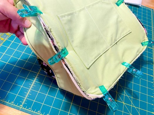 A hand holds the edge of a purse that is inside out. Large green clips hold the purse's layers together before sewing. A green cutting mat is visible in the background.