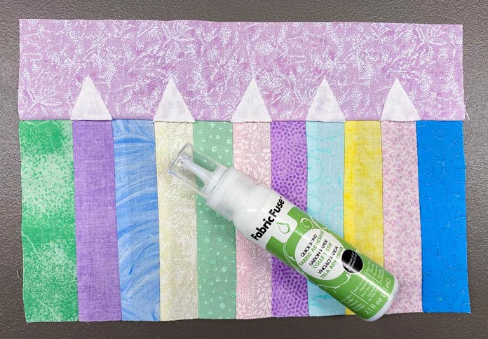 A bottle of Fabric Fuse by HeatnBond Quickbond Fabric Adhesive lying on top of the pastel-colored sewn piece of fabric with the white triangles as pencil tips