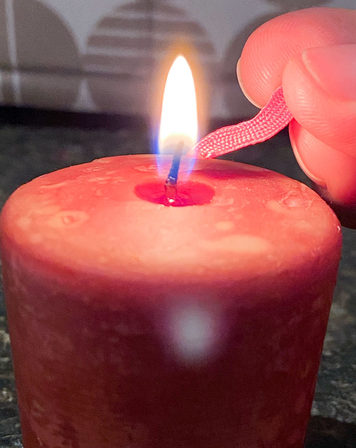 Fingers hold the end of a piece of pink rick rack to the bottom of the flame of a red candle flame. Brother NQ900 sewing machine, UNIQUE quilting Clever Clips, UNIQUE Medium Rick Rack