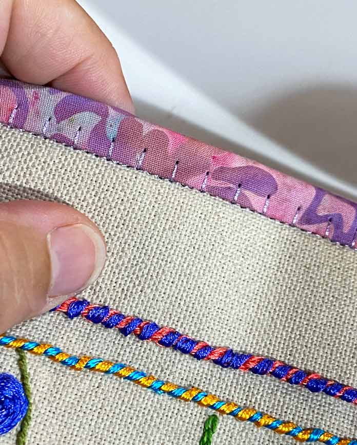 A closeup photo showing a hand holding the edge of tan embroidered fabric needle roll, with stitching on the binding prominently displayed.