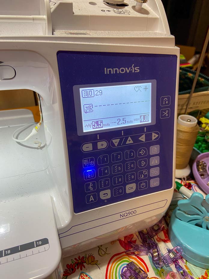 The blue and white Brother NQ900 sewing machine sits on top of a multicolored mat, and the screen is backlit and shows that stitch 29 is selected.