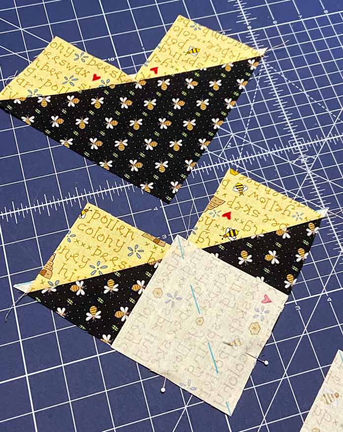 Two pieced black and yellow units for the flying geese block are shown on the blue cutting mat. One of the units has another marked square of yellow fabric pinned to it; OLFA Double Sided Cutting Mat (navy blue)