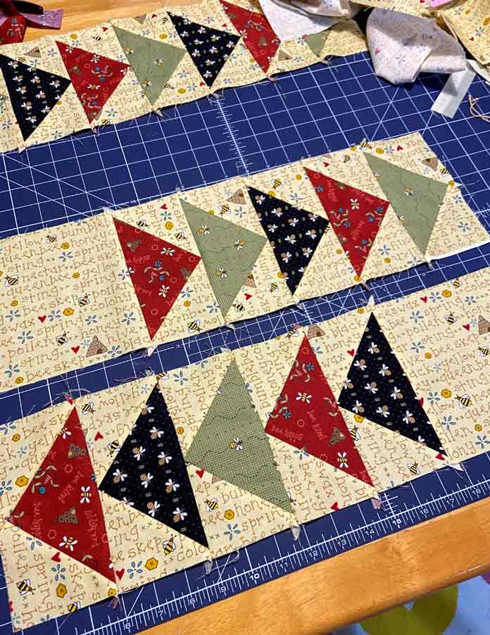 Two rows of five flying geese blocks in red, black, green and yellow lay on a dark blue cutting mat. One end of each of the borders has a yellow square of fabric sewn to it. A third border and scraps of fabric can be seen in the background; OLFA Double Sided Cutting Mat (navy blue)