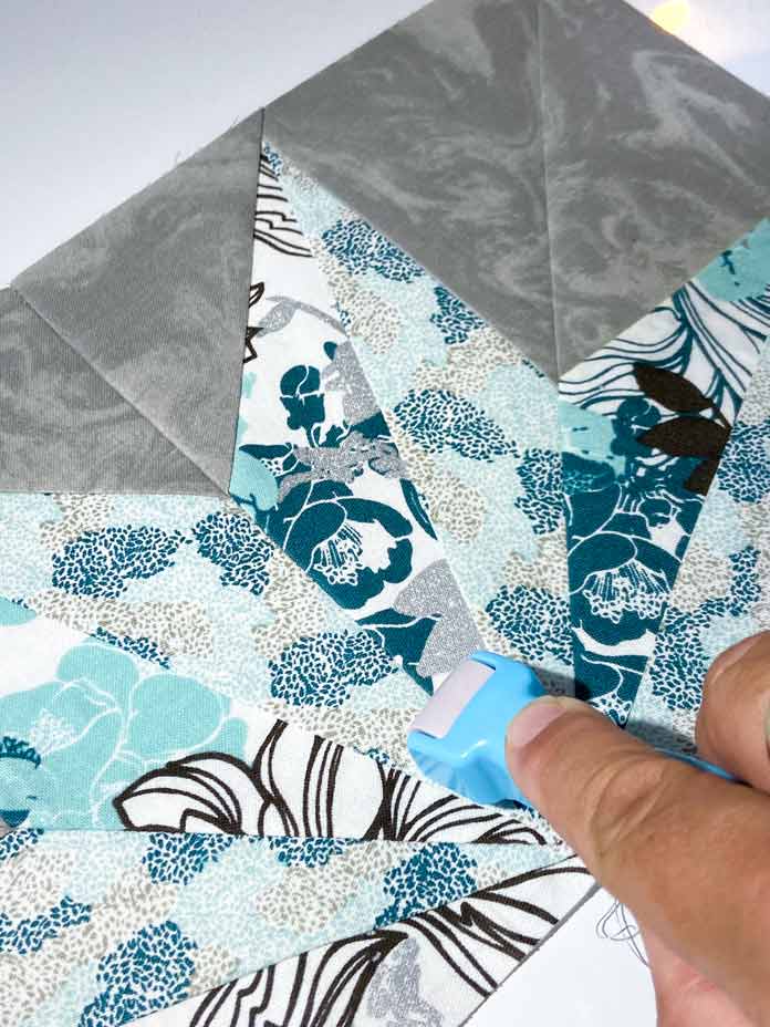 A hand uses a blue-handled Clover Roll & Press to flatten out the seam of the paper-pieced quilt block unit. StitchnSew EZ-Print Quilt Block Sheets, clover roll & Press, Clover Ball Point Awl, Fairfield Crafter’s Choice Pillow Form, Omnigrid 6"x 24" Ruler, OLFA 45mm Deluxe Ergonomic Handle Rotary Cutter, UNIQUE Double Sided cutting Mat