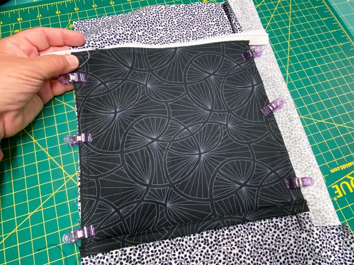 A black pocket with a white zipper is clipped to a larger piece of black and white fabric with purple clips. Everything lies on top of a green cutting board.