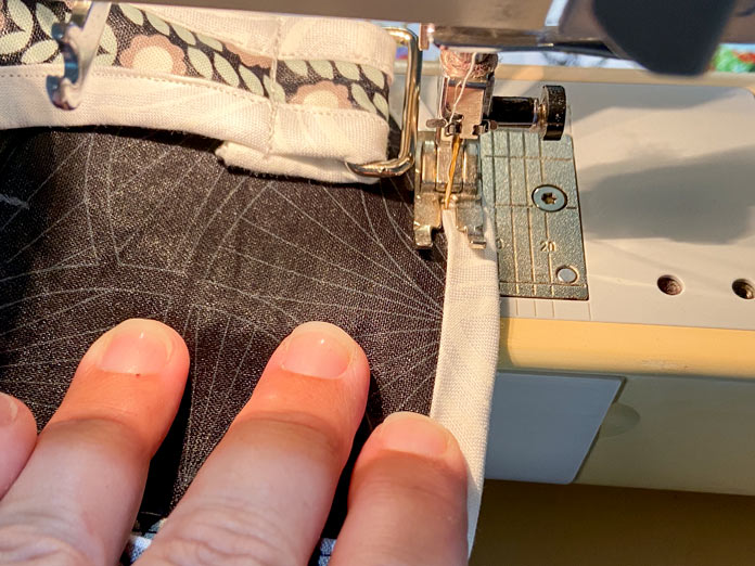 A hand feeds a black bag with white binding through a sewing machine so that the folded edge of the binding is topstitched.