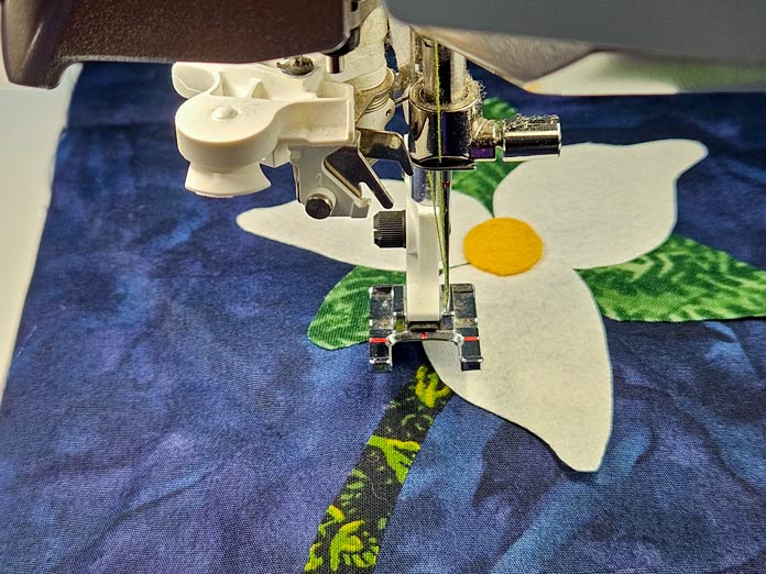 A white and green flower on blue fabric under a metal presser foot; Husqvarna VIKING DESIGNER EPIC 3