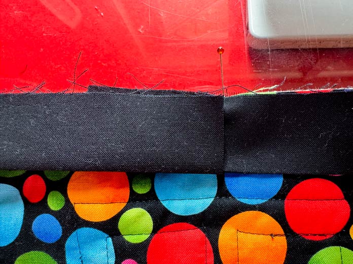 The start and end of a black binding strip, sewn to a quilt backing of black fabric with brightly colored circles on it, are laid on top of each other with a pin marking the position where the top strip overlaps and has been cut. A red sewing table is shown in the background. Hemline Gold Multi-Use Craft Bag (28.5cm x 36.5cm x 17.8cm), Hemline Gold Sewing Kit, Hemline Gold Quilters 4-in-1 Multi-Mat (30cm x 24cm), Hemline Gold Quilters Clips (Pack of 30), Hemline Gold Retractable Tape Measure - 150cm/60, oliso M3Pro Project Iron – Coral, OLFA 45mm RTY-2/DX/MAG Ergonomic Rotary Cutter – Magenta, OLFA RM-MG - 24″ x 36″ Double Sided Rotary Mat, UNIQUE Sewing Tools Holder, OLFA QR-6x12 - 6″ x 12″ Frosted Acrylic Ruler, OLFA QR-6x24 - 6″ x 24″ Frosted Acrylic Ruler, Fairfield Quilter's 80/20 Quilt Batting - 229 x 274cm (90″ x 108″) 