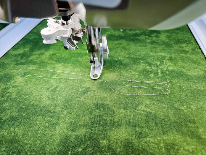 A machine embroidery foot stitching the shape of a fork with white thread on green fabric. Husqvarna VIKING DESIGNER EPIC 3 sewing and embroidery machine, Husqvarna VIKING 6in/15.2cm Applique Scissor, Husqvarna VIKING Open Toe Presser Foot for IDF System, Husqvarna VIKING Mega Turnable Hoop 450 mm x 450 mm, Husqvarna VIKING Tear-a-Way Extra Wide Stabilizer 24" x 10 Yards, Husqvarna VIKING Cut-a-Way Extra Wide Stabilizer 24” x 10 Yards, SULKY Slimline Metallic Dream Assortment