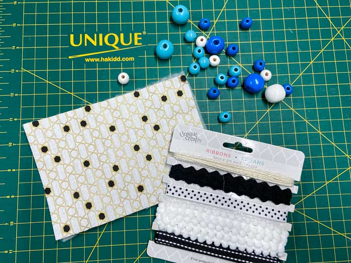 A package of black and white ribbons, blue and white wooden beads, and a white, gold, and black piece of fabric are shown sitting on a green cutting mat. OLFA CHN-1 - Chenille / Textile Cutter, UNIQUE QUILTING Stitch-N-Steam - 157 x 45cm (62″ x 18″), FABRIC CREATIONS Fabric Bundle (5pcs) - Metallic Gold - 45 x 53cm (18in x 21in), UNIQUE SEWING Self-Gripping Fastener Strip - 25cm (10″) - White, UNIQUE Webbing 25mm x 10m (1″ x 11yd) - White, an ELAN Trench Buckle - 45mm (13⁄4″) - Brown, UNIQUE CREATIV Trim Pack - Black & White, UNIQUE QUILTING Clever Clips Small - 12 pcs, CLOVER 7807 - Hold it Precision Stiletto, UNIQUE SEWING Sewing Tools Holder, UNIQUE QUILTING Wool Pressing Mat - 18″ x 24″ - Grey, OLISO M2Pro Mini Project Iron with Solemate, UNIQUE QUILTING Clever Clips Large - 12pcs, UNIQUE SEWING Invisible Magnetic Snaps - Sew-On - 12mm (1⁄2″) - 2 sets, COSTUMAKERS Activewear One Way Separating Zipper 23cm (9″) - White - 1760, UNIQUE SEWING Double Adjust Parachute Buckle - Plastic - 25mm (1″) - White, free tutorial