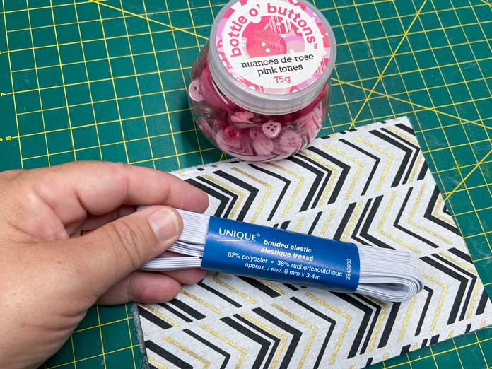 A hand holds a blue package of braided elastic beside a bottle of pink buttons. In the background, a piece of fabric with a black and gold chevron pattern sits on a green cutting mat. UNIQUE SEWING Self-Gripping Fastener Strip - 25cm (10″) - White, UNIQUE Webbing 25mm x 10m (1″ x 11yd) - White, ELAN Trench Buckle - 45mm (13⁄4″) - Brown, UNIQUE CREATIV Trim Pack - Black & White, UNIQUE SEWING Invisible Magnetic Snaps - Sew-On - 12mm (1⁄2″) - 2 sets,  SOFTKUT Pinking Shears (Scissors) - 81⁄2″ (21.6cm), COSTUMAKERS General Purpose Closed End Zipper 20cm (8″) - White - 1700, UNIQUE CREATIV Trim Pack - Red & Green, OLFA RTY-2/DX - Deluxe Ergonomic Handle Rotary Cutter 45mm, UNIQUE QUILTING Clever Clips Small - 12 pcs, UNIQUE SEWING Double Adjust Parachute Buckle - Plastic - 25mm (1″) - White, CRAFTING ESSENTIALS Bottle of Buttons - Pink Tones - 75g (2.6oz), UNIQUE Braided Elastic 3mm x 3.7m - White, free tutorial 