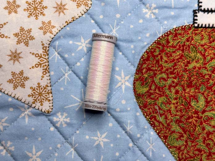 A spool of white Gütermann metallic thread sits on top of a quilted tabled runner; Gütermann Dekor Metallic Thread 200m – Gold, Gütermann Dekor Metallic Thread 200m - Col. 41, Gütermann Dekor Metallic Thread 200m – Red, Gütermann Dekor Metallic Thread 200m - Christmas Green, Gütermann Dekor Metallic Thread 200m - Col. 1005, Gütermann Denim Thread 100m – Black, UNIQUE Baby Rick Rack 3mm x 3.7m – Scarlet, UNIQUE Medium Rick Rack 6mm x 2.3m - Emerald Green, UNIQUE Jumbo Rick Rack 10mm x 2.3m - Metallic Gold, SCHMETZ #1743 Metallic Needles Carded - 80/12 - 5 count, SCHMETZ #1752 Metallic Needles Carded - 90/14 - 5 count, LDH 8½“ Craft Scissors – Purple, HeatnBond Lite Iron-On Adhesive - 43cm x 30m (17″ x 32 ¾yds) bolt, HeatnBond Stitch n Sew Tear-Away Stabilizer - 50.8cm x 22.8m (20″ x 25yds), oliso M3Pro Project Iron – Coral, OLFA 45mm RTY-2/DX/MAG Ergonomic Rotary Cutter – Magenta, UNIQUE Quilting Wool Pressing Mat - 18″ x 24″ - Grey