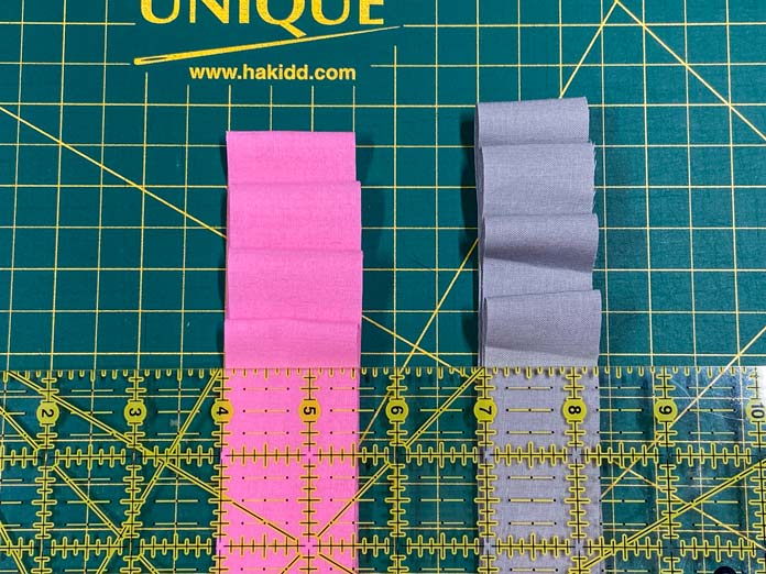 A stack of 1½” wide pink strips and a stack of 1¼” wide gray strips sit on top of a green cutting mat. A rotary cutter ruler with a yellow grid sits on top and shows the width of the strips.