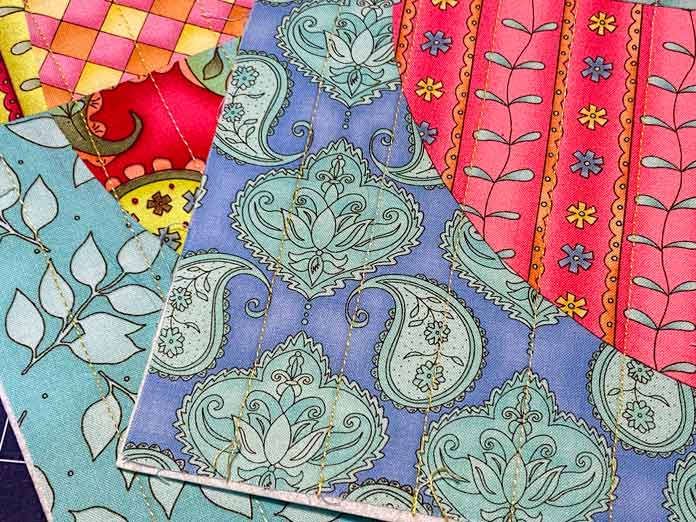 Two multicolored quilt blocks that have been machine quilted in straight lines using yellow thread sit on top of each other. 