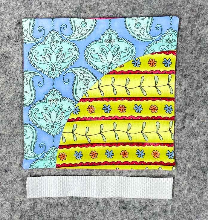 A multicolored quilt block sits on a grey pressing mat. A short piece of white webbing sits below it.