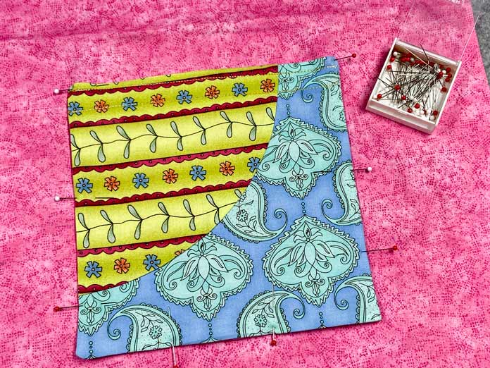 A multicolored pocket has been pinned on three sides to a pink lining fabric. A small box of white and red-headed pins sits open beside the pocket.