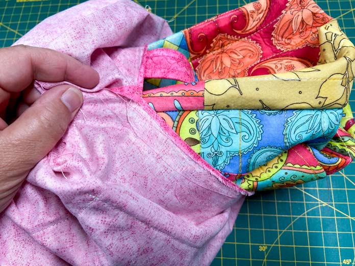 A hand holds the lining of a tote bag. The outside of the tote bag is being pulled out through a hole in the bottom of the lining. A green cutting mat can be seen in the background.