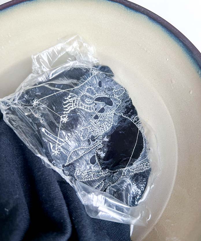 The embroidered area of the shirt is visible, showing a detailed white line-art design stitched onto the fabric. The layer of water-soluble film topper is beginning to dissolve and peel away in soft folds, exposing the embroidery underneath. 
