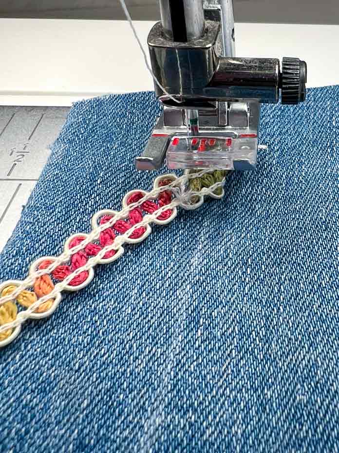 Close-up view of the PFAFF creative icon 2 sewing machine performing a Single Ribbon Stitch. The ribbon is manually folded over the previous stitch section, ready for the next segment. The decorative ribbon is aligned in front of the needle, and the 2A Fancy Stitch Foot is attached. The denim fabric shows the partially completed ribbon stitch design with precise positioning.