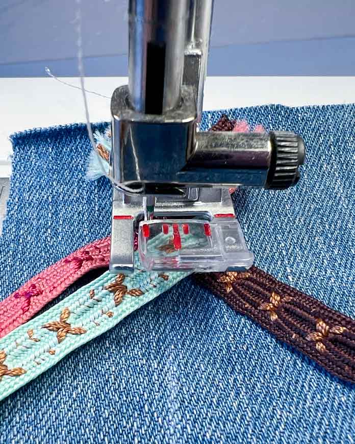 Close-up of the 2A Fancy Stitch Foot on the PFAFF creative icon 2, with brown, teal and pink ribbons positioned on denim. The third (teal) ribbon is folded over the first (brown) and positioned in front of the needle and parallel to the second (pink) ribbon to complete one cycle of the fold sequence, ready for the next round of stitching. This step shows the controlled placement before pressing Start to continue the Triple Ribbon Stitch.