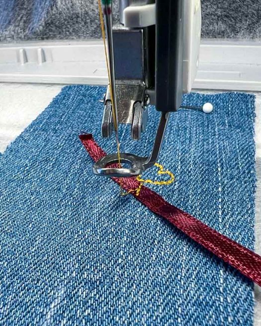 Close-up of the ribbon placement step during embroidery on the PFAFF creative icon 2. A 4 mm burgundy satin ribbon is positioned over a yellow placement stitch on medium-weight denim fabric. The presser foot is lowered, and the needle is aligned just above the ribbon, ready to continue stitching. The PFAFF 6D Dynamic Spring Foot is in use, and the design is hooped in the Elite Hoop 260 x 200 mm. The fabric is stabilized with white tear-away backing for optimal support.