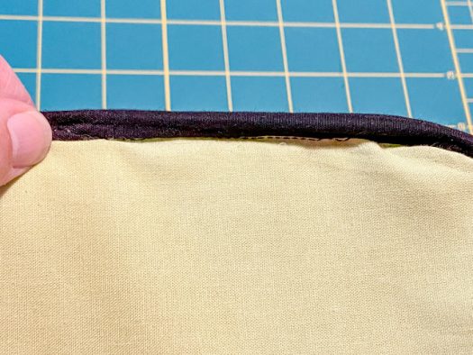 A hand holds the edge of a purse that is inside out to show the black bias tape that has been sewn over the inside seams. A green cutting mat is visible in the background.