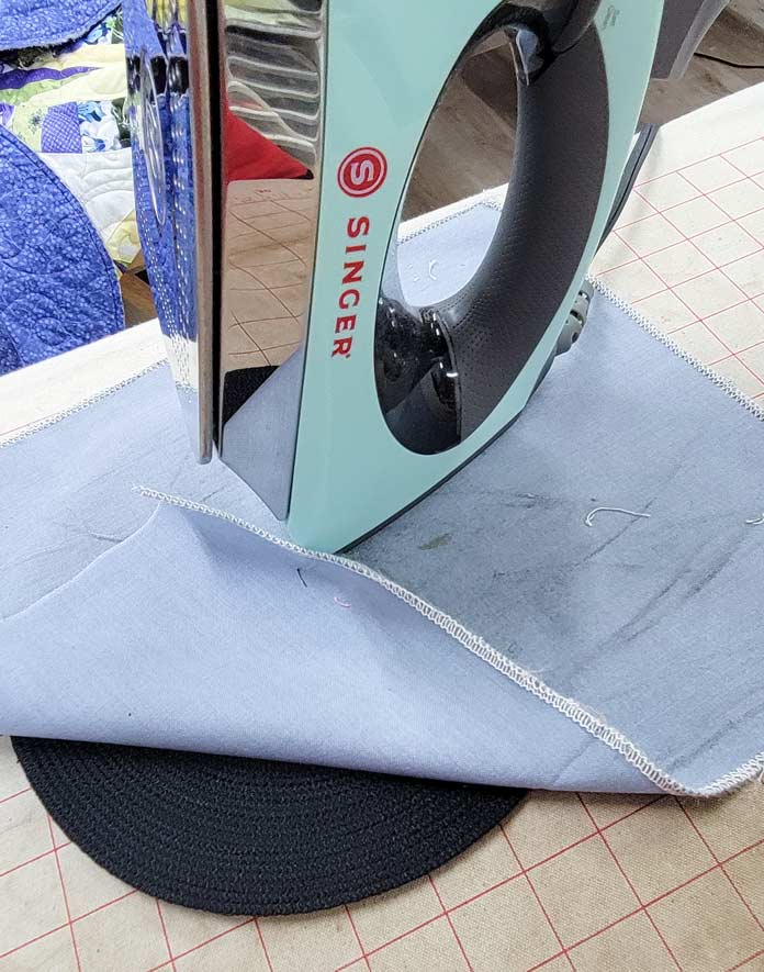 A black circle of stitched cording under a press cloth on an ironing board with a green iron; Husqvarna Viking Designer Ruby 90 sewing and embroidery machine, Singer Ironing and Crafting Station, Singer Steam Craft Plus Iron, Inspira Aqua Magic Plus Stabilizer