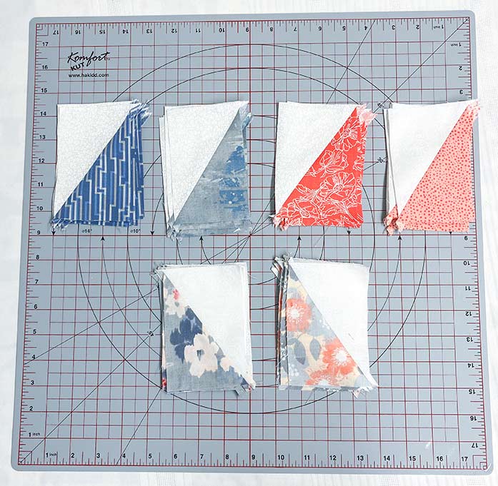 Half Rectangle Triangle units pressed open and sorted into six piles. SCHMETZ Quilting needles, UNIQUE Sewing Fast Fade Fabric Markers, Omnigrid Rulers, OLFA Rotary Cutter, Komfort Kut Rotating Cutting mat, Fabric Creations Cotton Fabric.