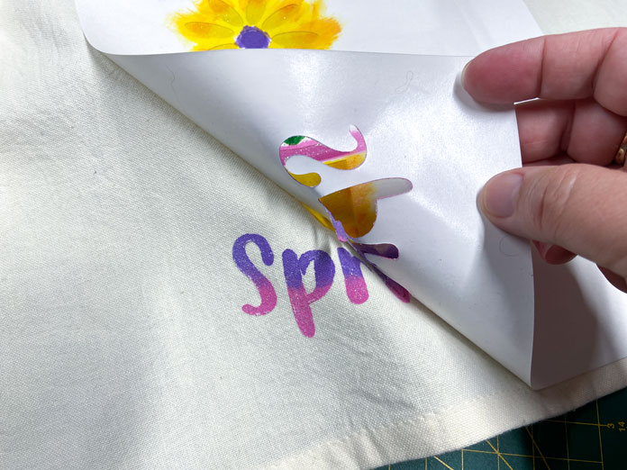 A hand is shown removing a white stencil from a beige tea towel. The pink and purple stenciled design can be seen to the left of the stencil; Sew Easy Freezer Paper for Quilting and Applique, Mont Marte Signature Fabric Paints, Odif OdiShine Glitter gel, Odif OdiShine Glitter Gel – Opal 65g
