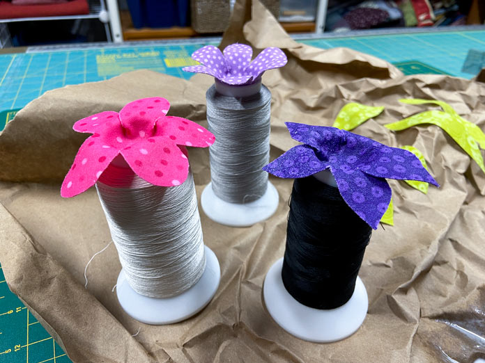 Pink, dark purple and light purple fabric flowers are shown sitting on the top of three large spools of thread. A crumpled brown paper and a few green fabric leaves can be seen in the background.