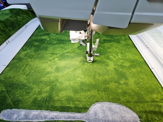 An embroidery presser foot on green fabric hooped in a gray machine embroidery hoop while stitching with green thread is in process. A gray applique spoon is towards the front of the hoop. Husqvarna VIKING DESIGNER EPIC 3 sewing and embroidery machine, Husqvarna VIKING 6in/15.2cm Applique Scissor, Husqvarna VIKING Open Toe Presser Foot for IDF System, Husqvarna VIKING Mega Turnable Hoop 450 mm x 450 mm, Husqvarna VIKING Tear-a-Way Extra Wide Stabilizer 24" x 10 Yards, Husqvarna VIKING Cut-a-Way Extra Wide Stabilizer 24” x 10 Yards, SULKY Slimline Metallic Dream Assortment